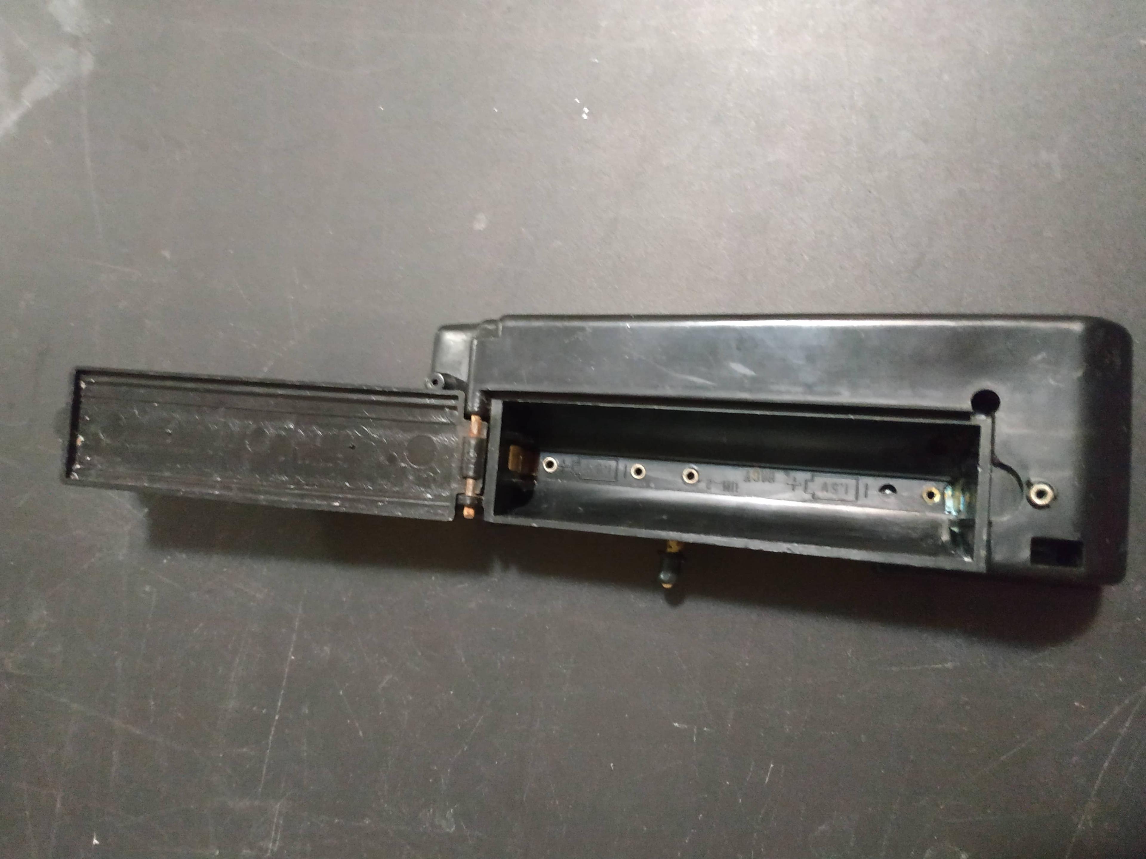 A black plastic part with a hinged battery compartment. There is noticeable blue corrosion in the corner
