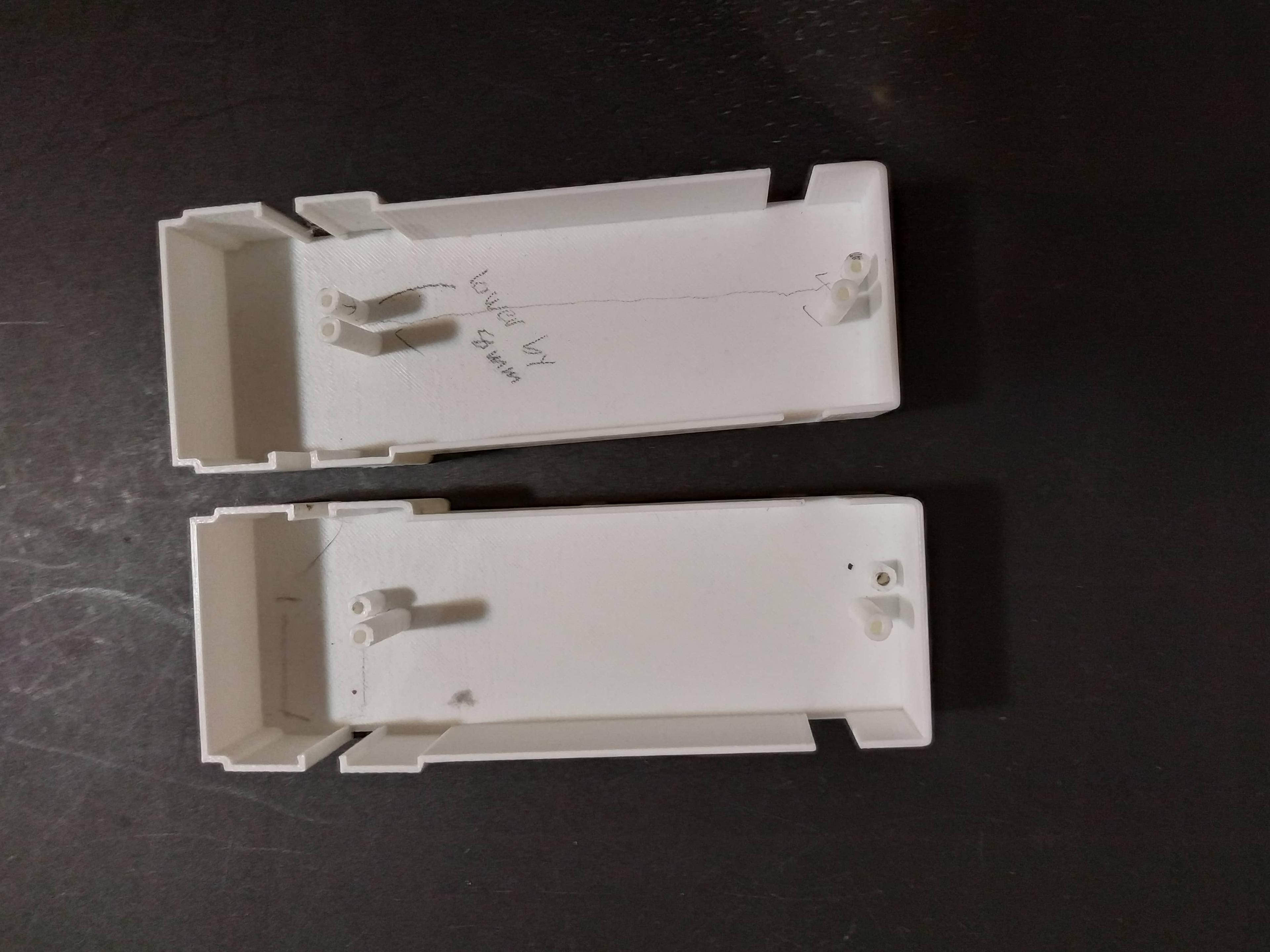 Two white plastic 3D models sit on a table, facing up. They have a 3D outline and extrusions for screw holes coming up from a flat base, and there are some pencil markings on them indicating proposed design changes