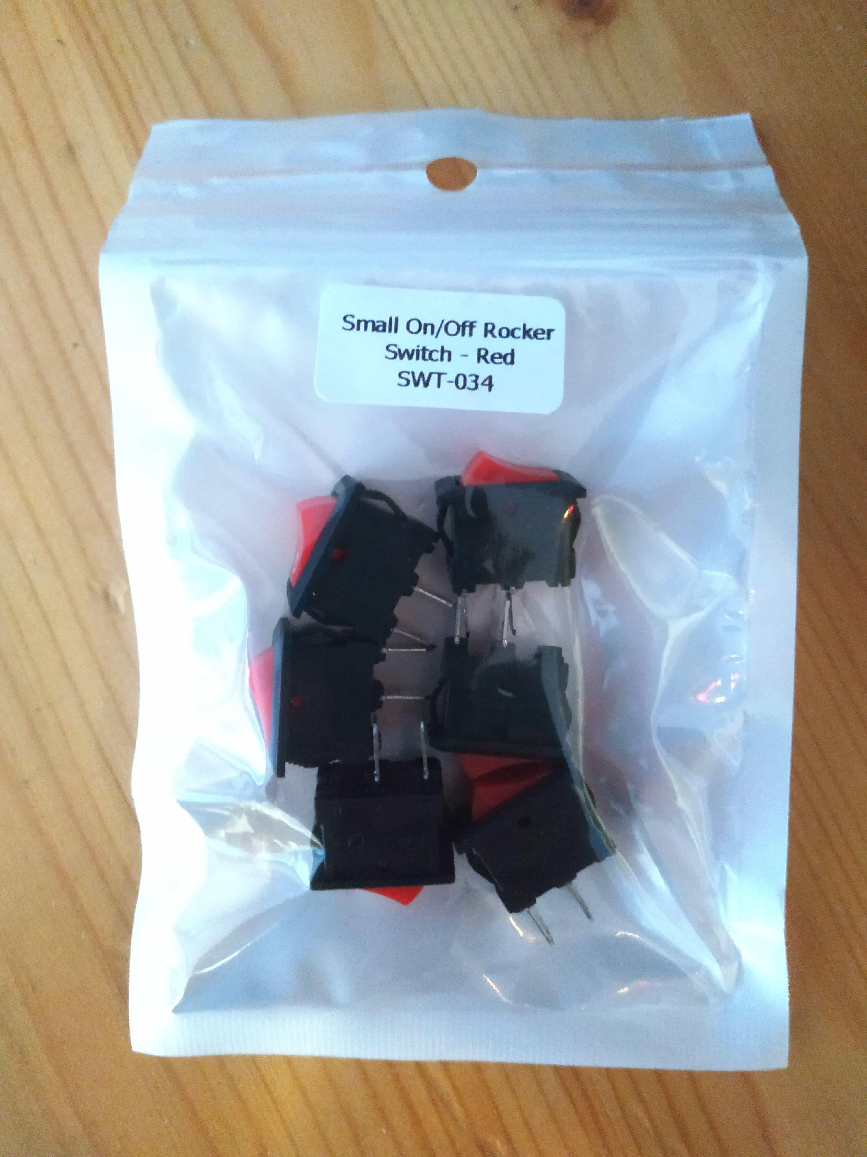 Six black rocker switches, with red on/off parts, lie in a plastic bag marked 'Small On/Off Rocker Switch - Red SWT-034'