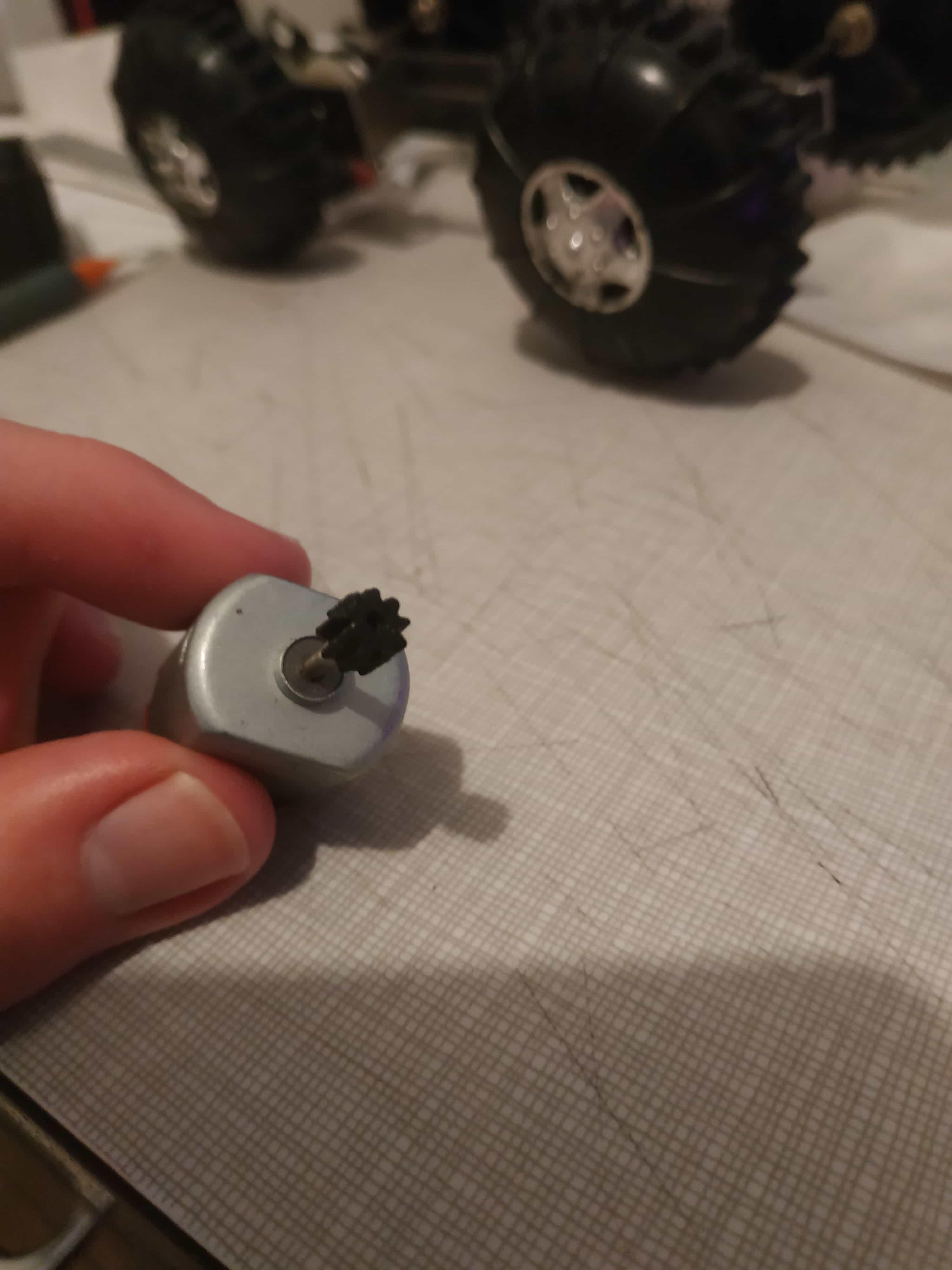 My hand holding up a small DC motor with a tiny plastic gear on it, the buggy is in the background