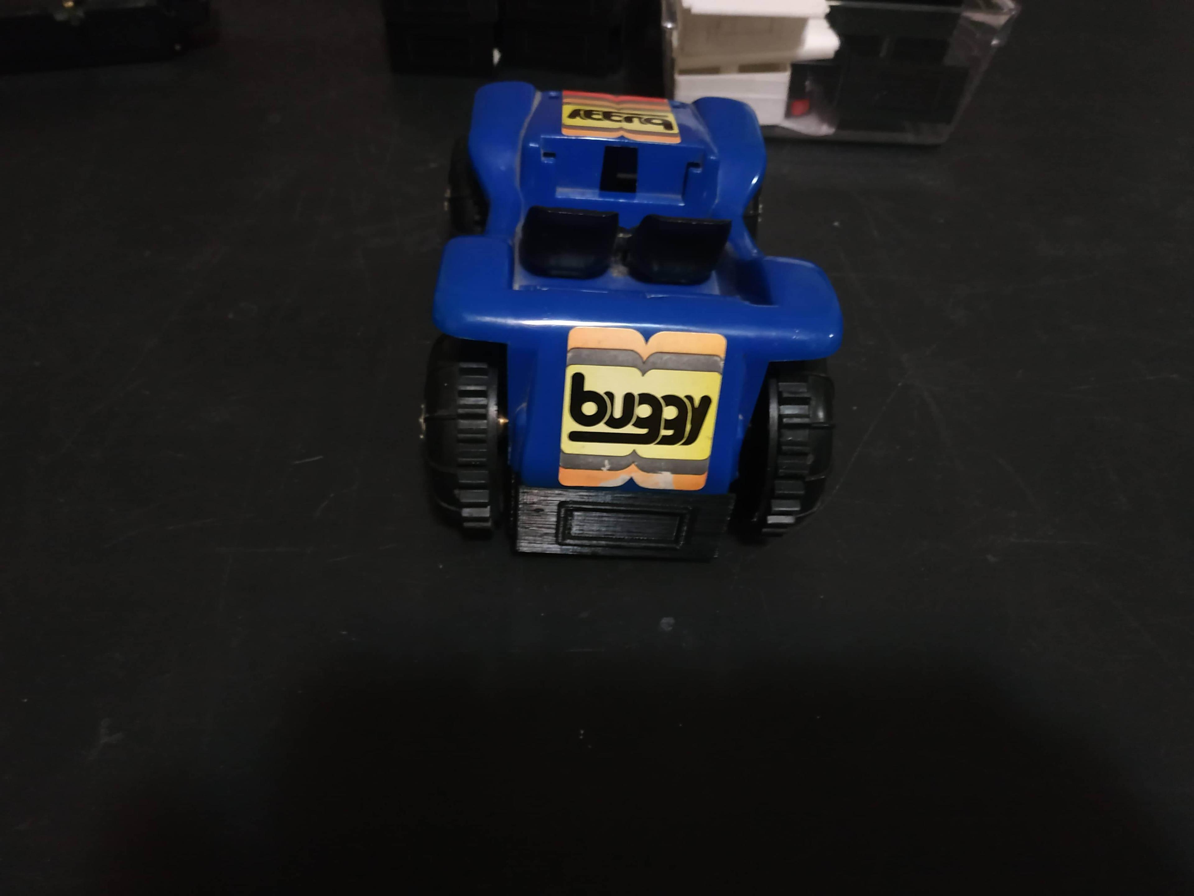 The back of the buggy. Its blue plastic top has a yellow and orange sticker that says 'buggy' on it. Below the top is the black 3D-printed base, with a shape that looks like a license plate