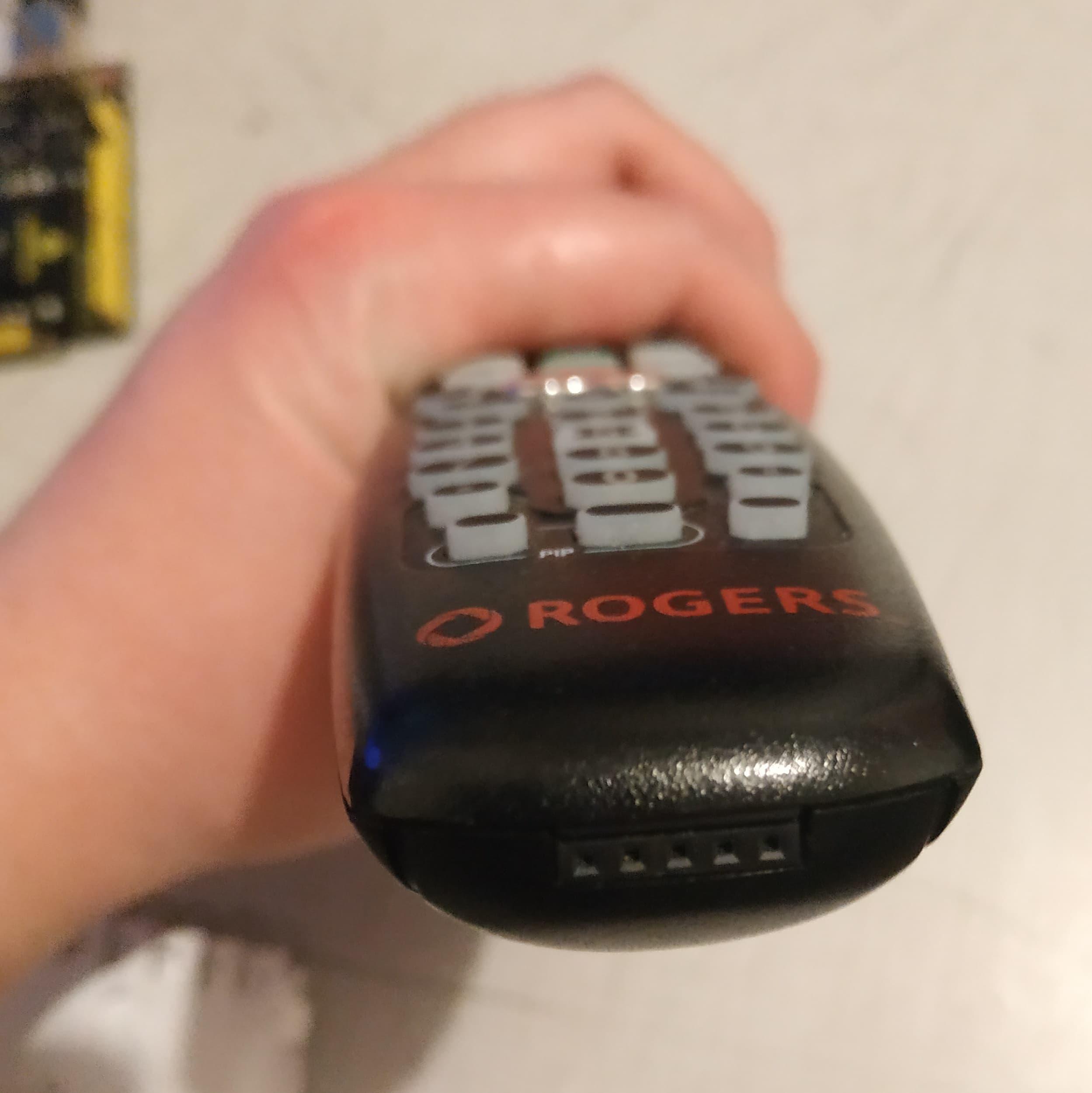 My hand holding a TV remote over a desk, with the bottom part towards the camera. The Rogers logo appears below the number pad. And below that, on the very bottom of the remote, is a black rectangular programming port