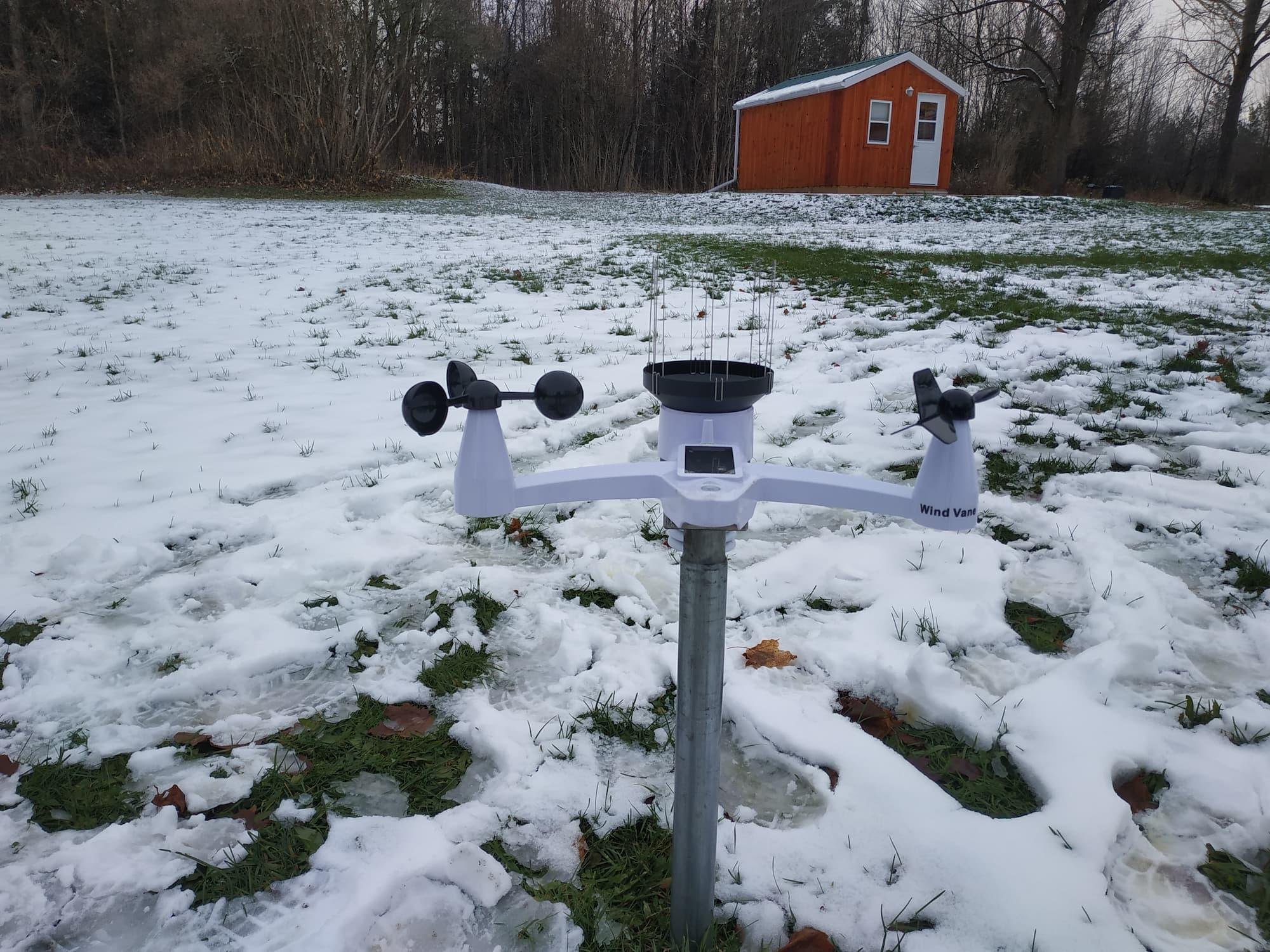 A white and black weather monitoring unit with an anemometer, rain collector, wind vane, solar panel, and other sensors is atop a short metal pole, with snow on the ground and my red office cabin in the background
