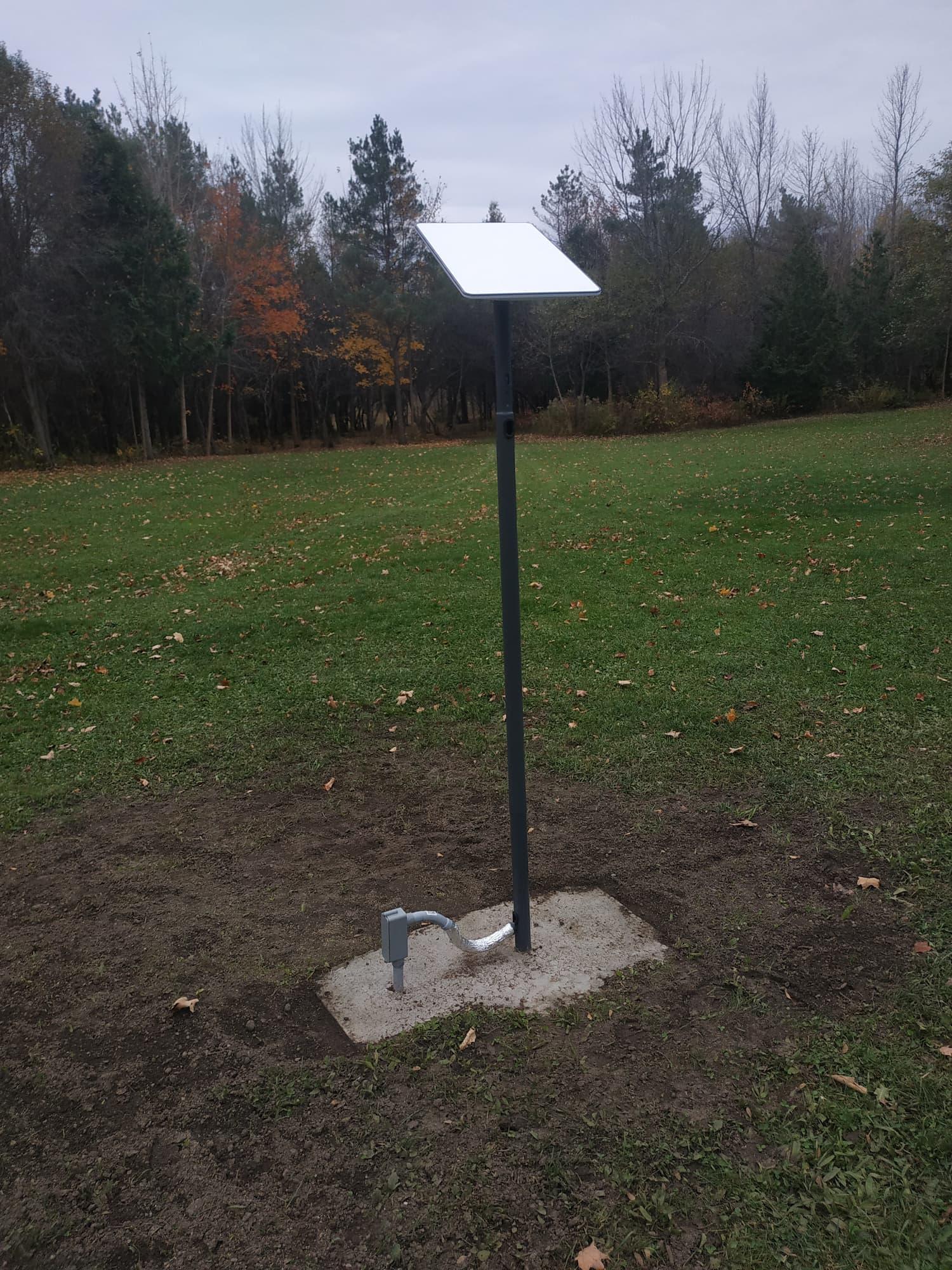 My white Starlink dish is positioned on a metal pole a few feet above the ground, with a cement pad below it. Part of a grey conduit is sticking up out of the ground, to which the wire is attached. The pole has been erected on a lawn