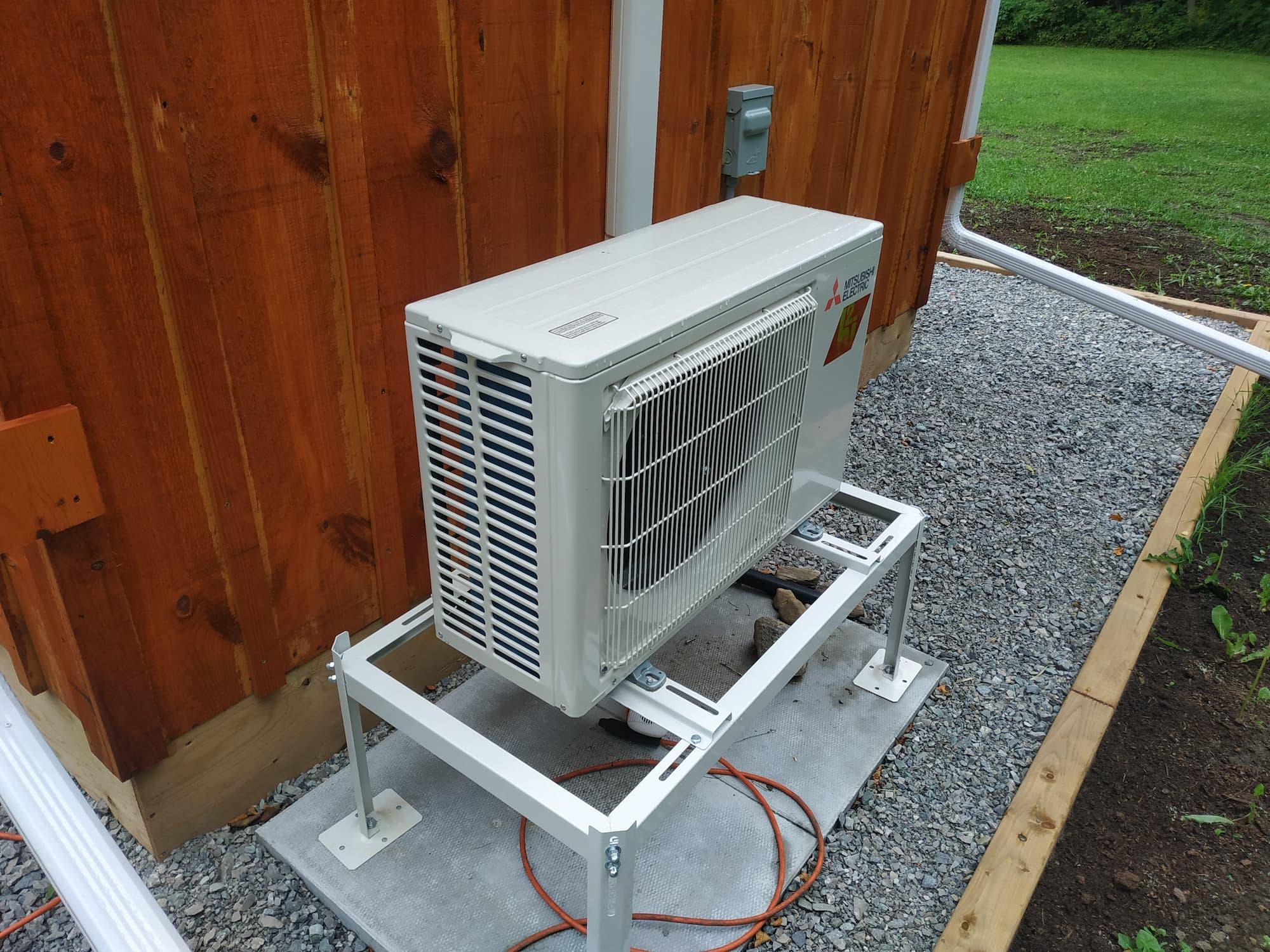 A small heat pump unit is seated on a metal stand, on top of a concrete slab, next to the back wall of my cabin