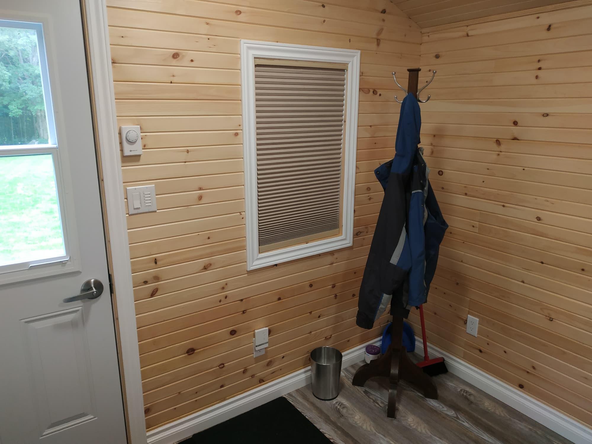 A coatrack sits in the corner, beside a broom and a small waste basket. A window with the shade pulled down is beside the coatrack, to the right of the door.