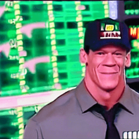 John Cena smiles, wearing a baseball cap, a grey shirt, and a black tie, in front of a green background that resembles a stock chart